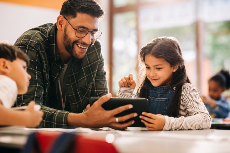 Educação socioemocional: como trabalhar emoções na escola? 3 Foto de estudante em sala de aula, ao lado de professor, fazendo atividade online em tablet sobre temas socioemocionais